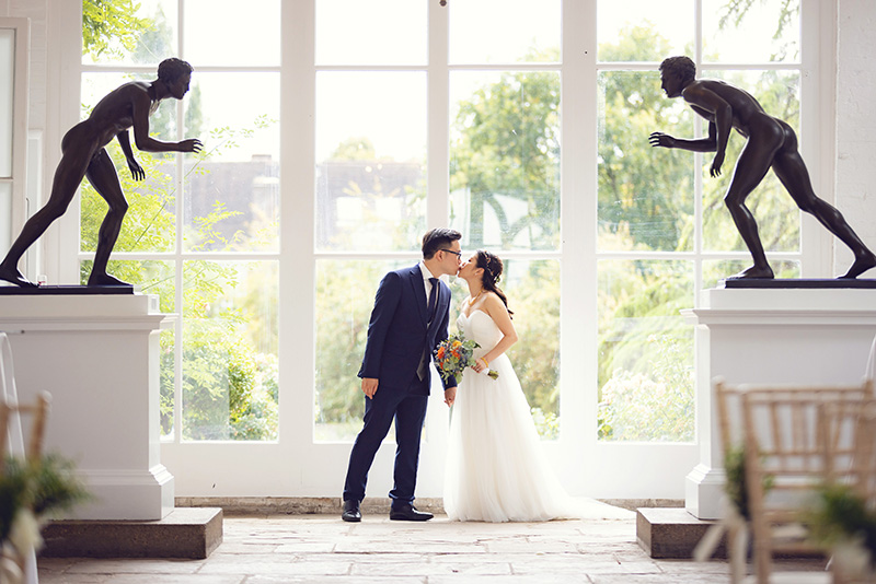 Bride and groom kissing at wedding ceremony in bright window-lit venue with statues.